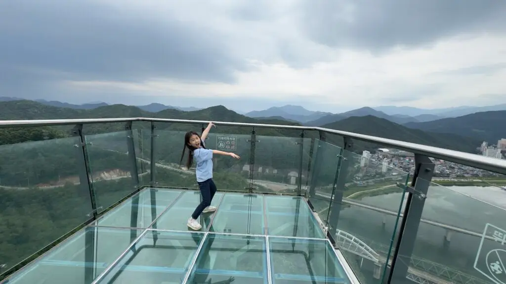 Young girl posing on the observation deck at 만천하스카이워크 in Danyang, South Korea, with scenic mountains and river in the background.