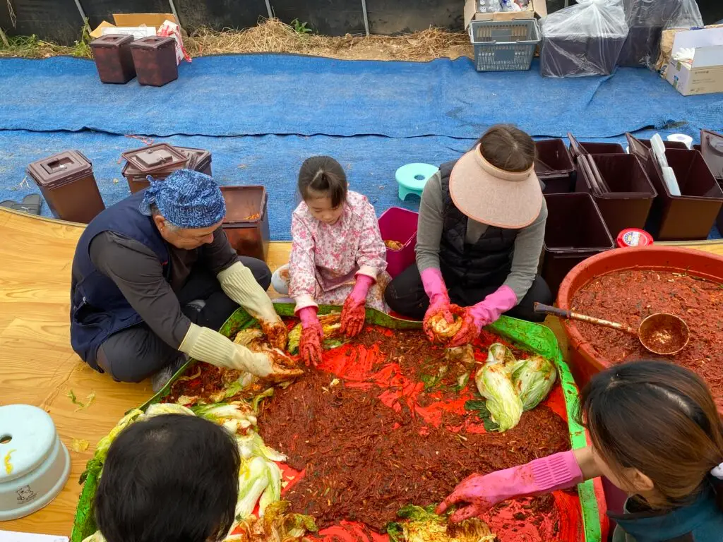Family gathered together making kimchi (traditional Korean kimjang), sitting around a large table covered with napa cabbage and red pepper seasoning.